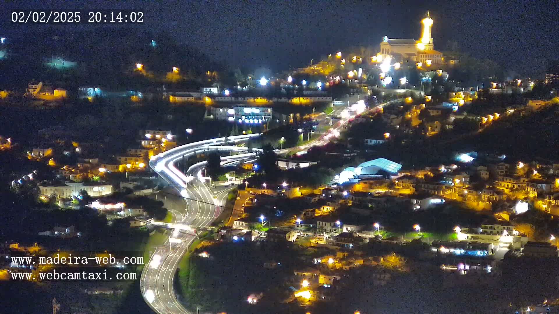 A nighttime aerial view of a town nestled on a hillside, illuminated by streetlights and building lights, with a highway curving through the landscape and a church atop the hill.