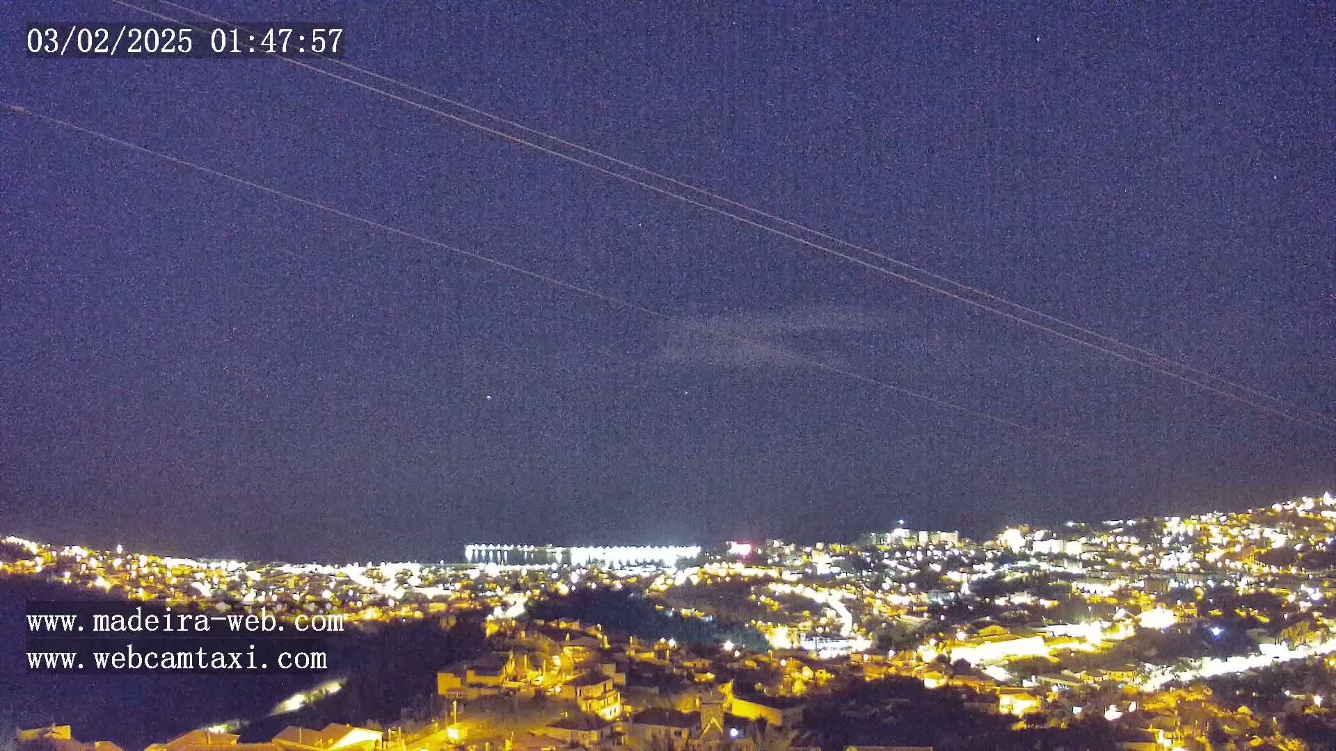 A nighttime view of a city illuminated by lights under a clear, dark sky with a few faint clouds and power lines visible.
