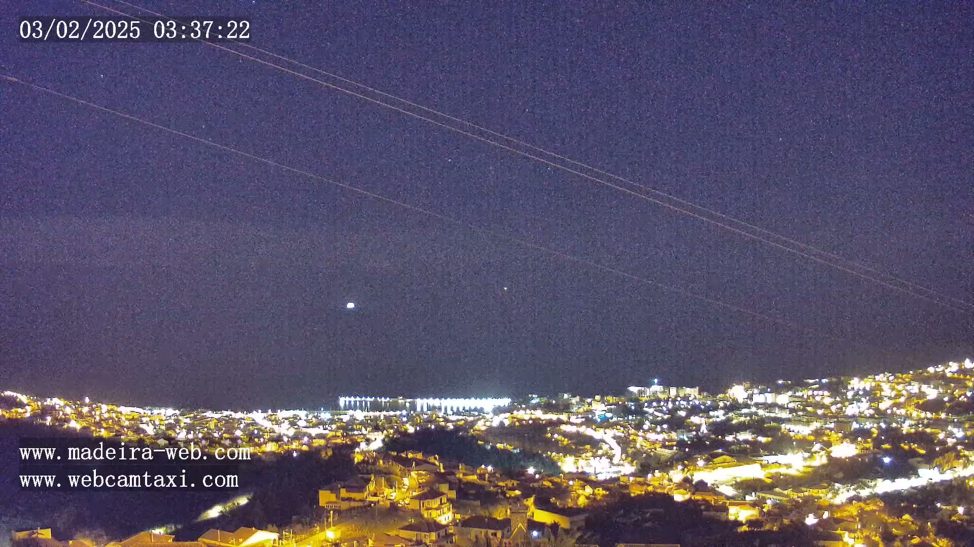A nighttime view of a city illuminated by lights under a clear, dark sky with several faint streaks across it.
