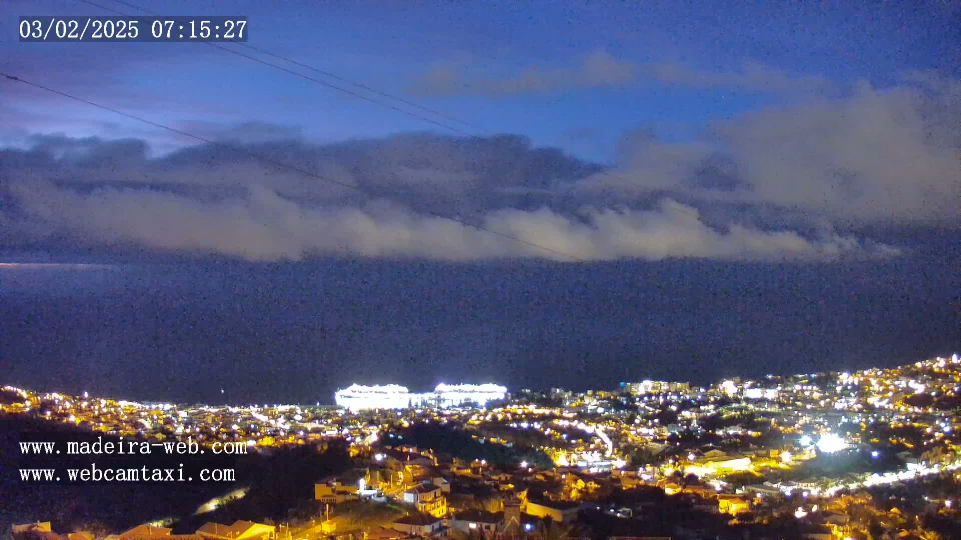 A nighttime coastal town with illuminated buildings is visible under a partly cloudy sky.