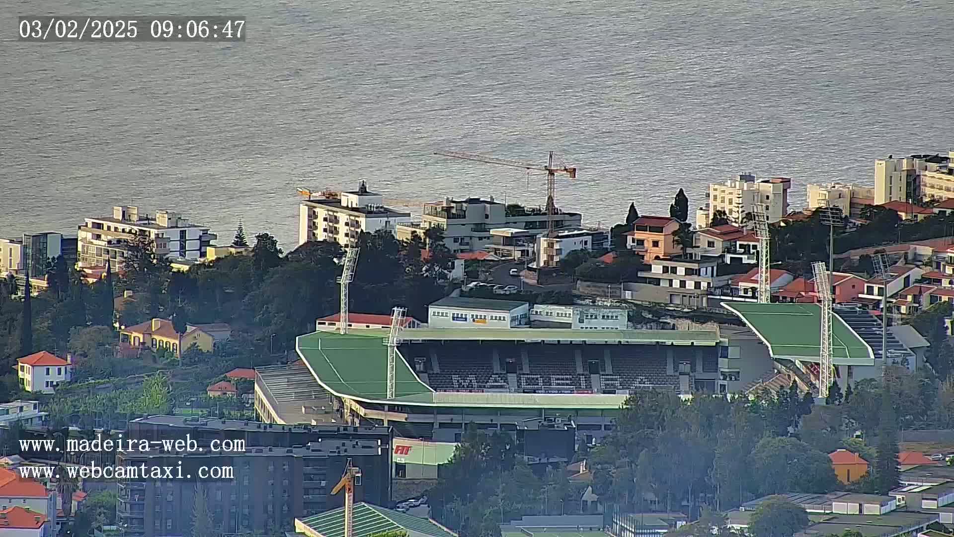 An aerial view shows a green stadium nestled among residential buildings overlooking a calm body of water under a partly cloudy sky.