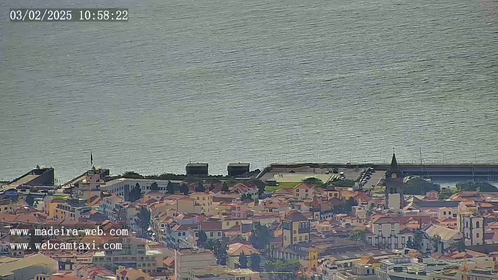A coastal town with predominantly low-rise red-roofed buildings extends to a calm, gray-blue sea under a clear sky.