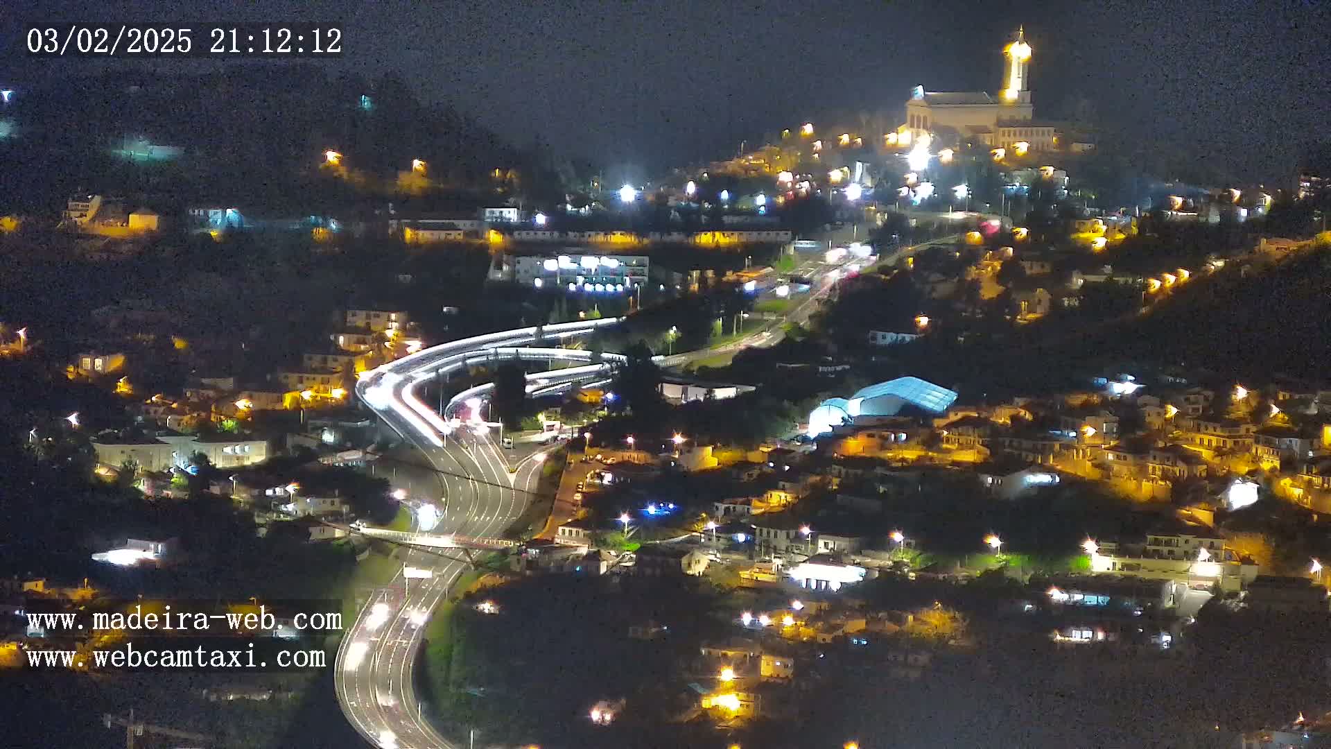 A nighttime aerial view of a town nestled on a hillside, with illuminated buildings, roads, and a church at its peak, under a clear, dark sky.