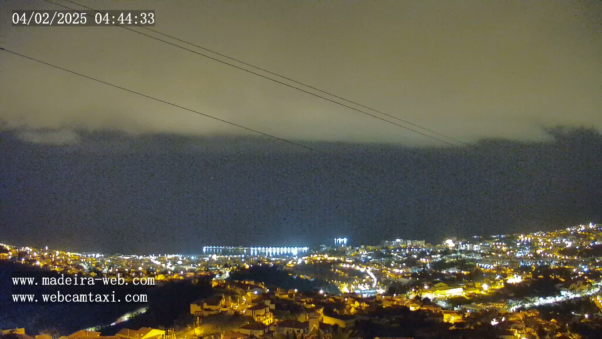 A nighttime view of a coastal town illuminated by lights, with a hazy or cloudy sky above.