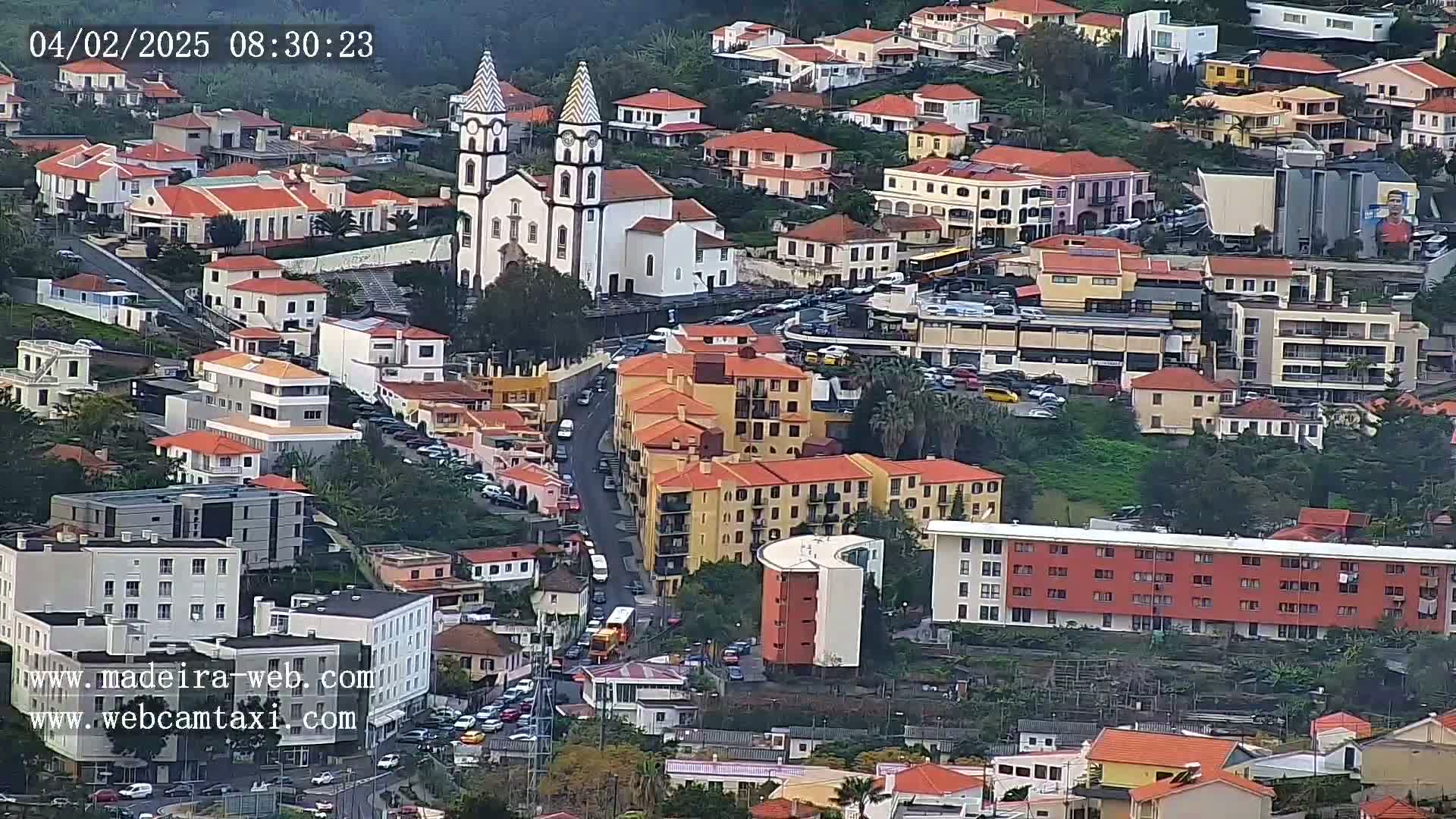 An aerial view shows a dense collection of buildings, including a church with twin towers, on a hillside under an overcast sky.