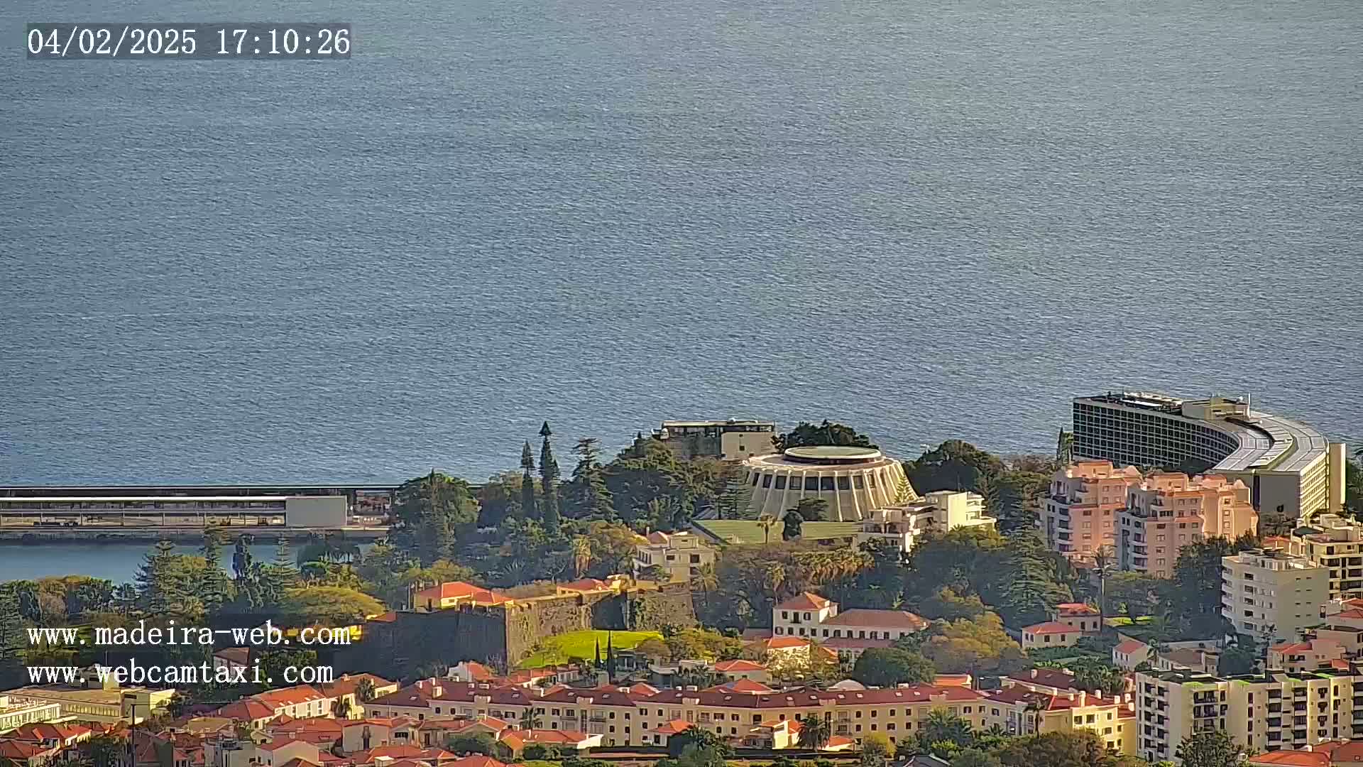 A coastal city with buildings of varying heights, including a large circular structure and a curved hotel, overlooks a calm blue ocean under a clear sky.