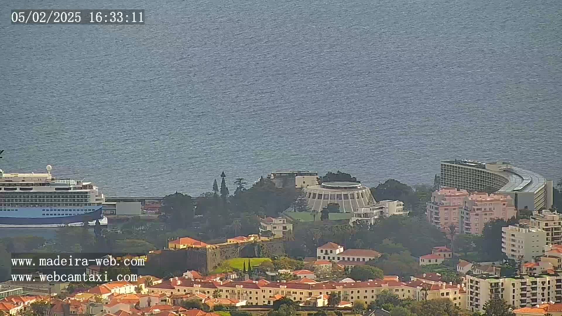 A cruise ship is docked near a coastal city with numerous buildings, including a large hotel, under a mostly clear sky.