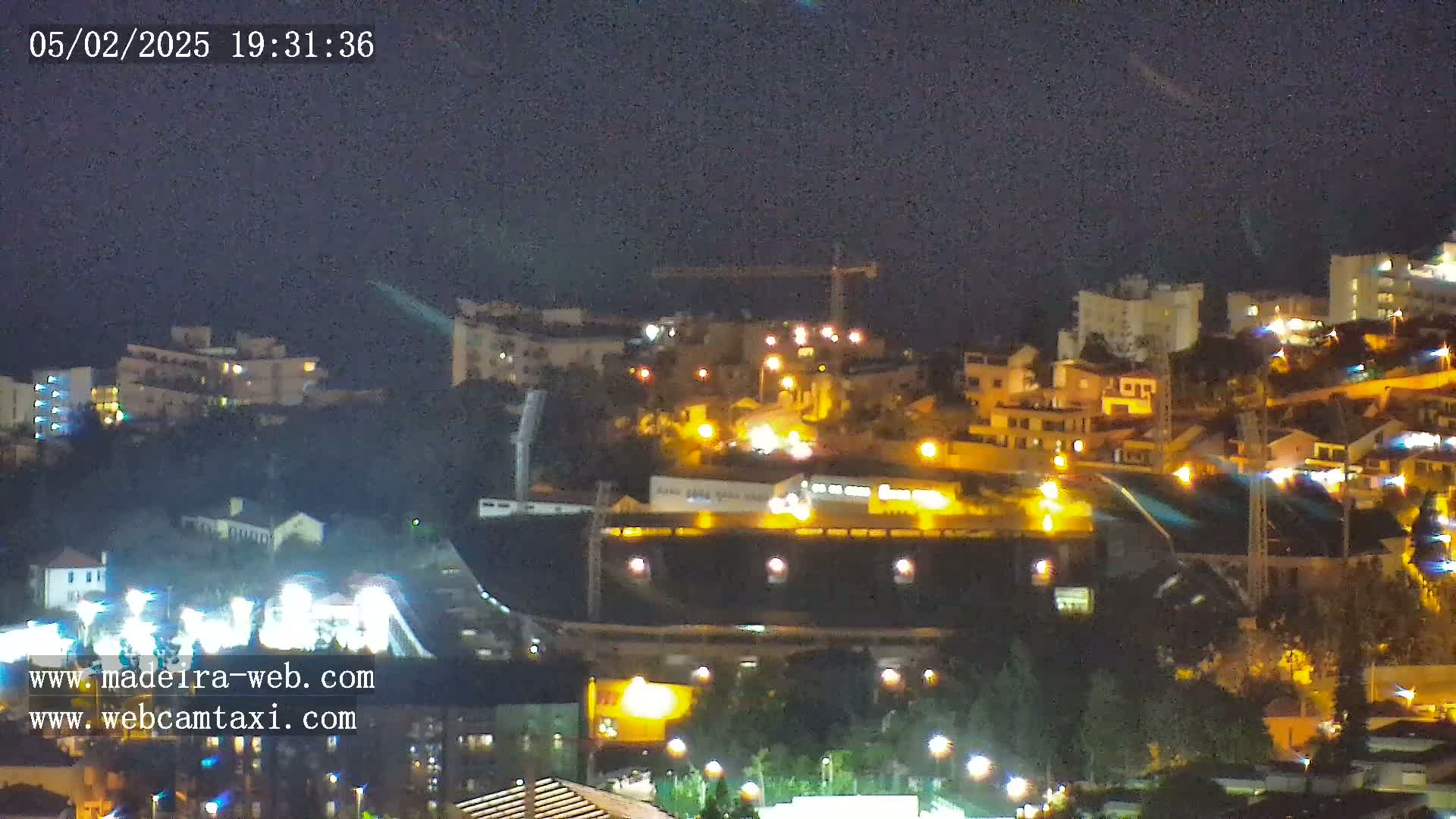 A nighttime view of a hillside town illuminated by numerous lights, featuring buildings, a stadium, and construction cranes under a dark sky.