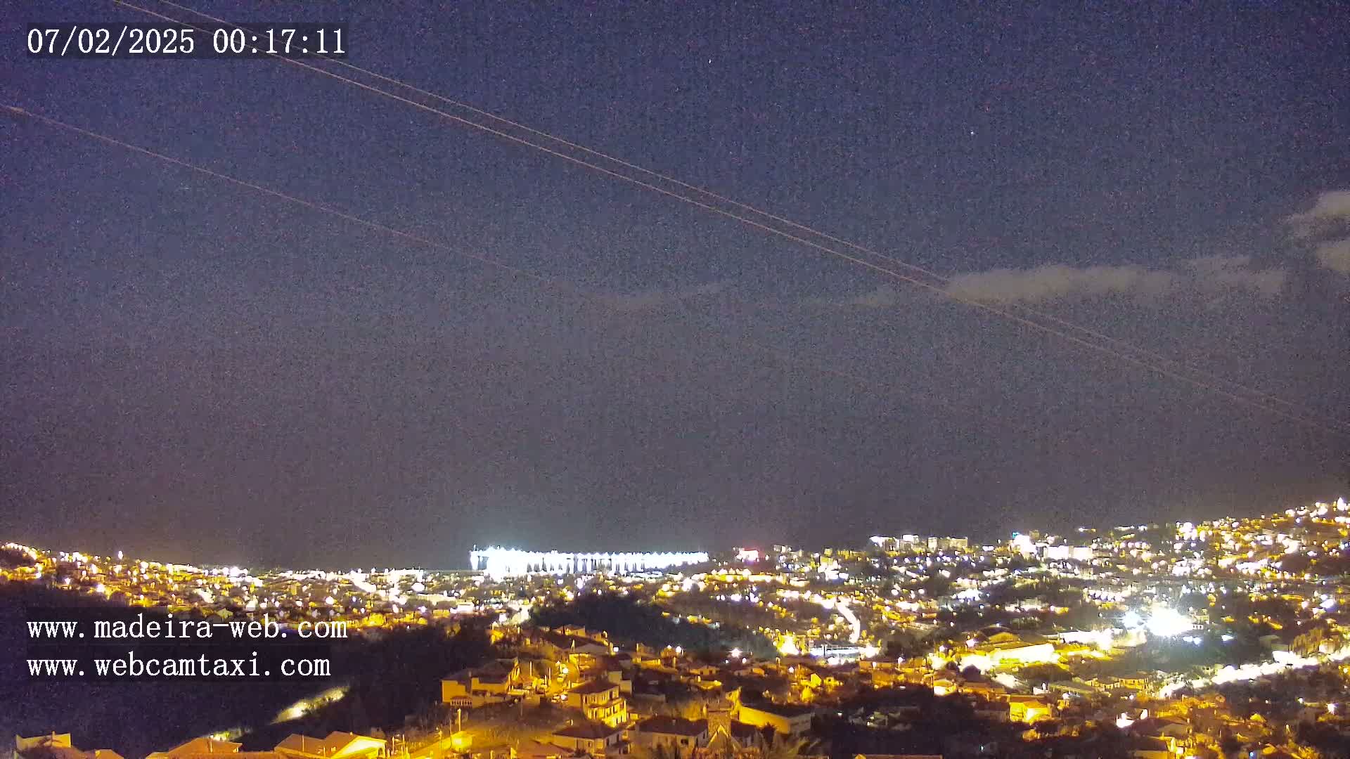 A nighttime long shot of a city illuminated with lights under a mostly clear, dark sky with a few thin clouds.