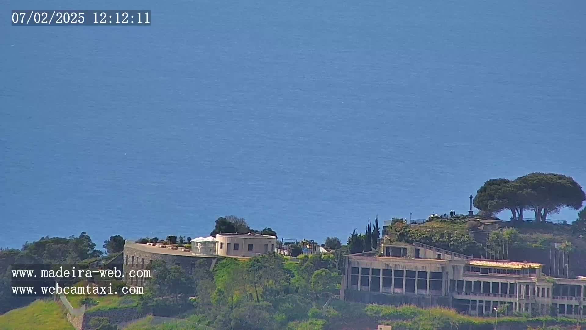 A coastal scene shows a large building complex overlooking a calm blue ocean under a clear sky.
