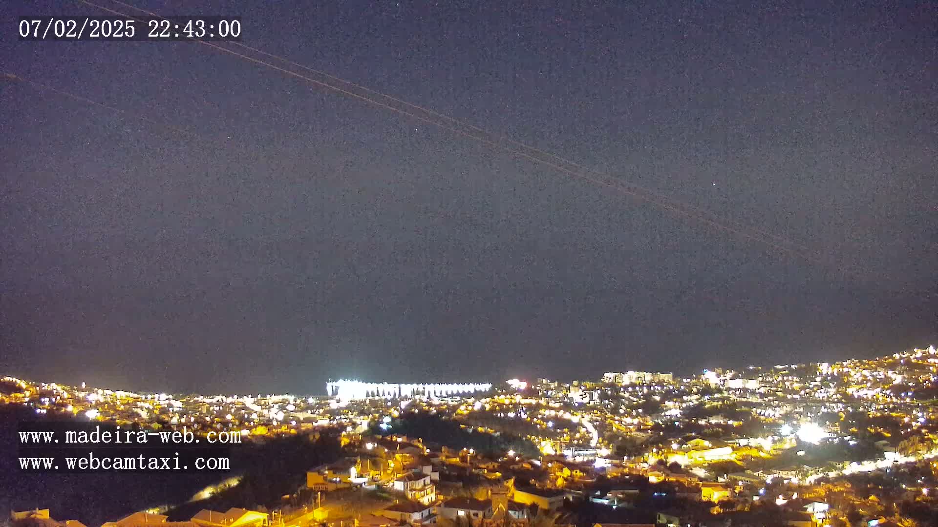 A nighttime, long shot of a city illuminated by lights, with a clear, dark sky showing a few stars and airplane contrails.
