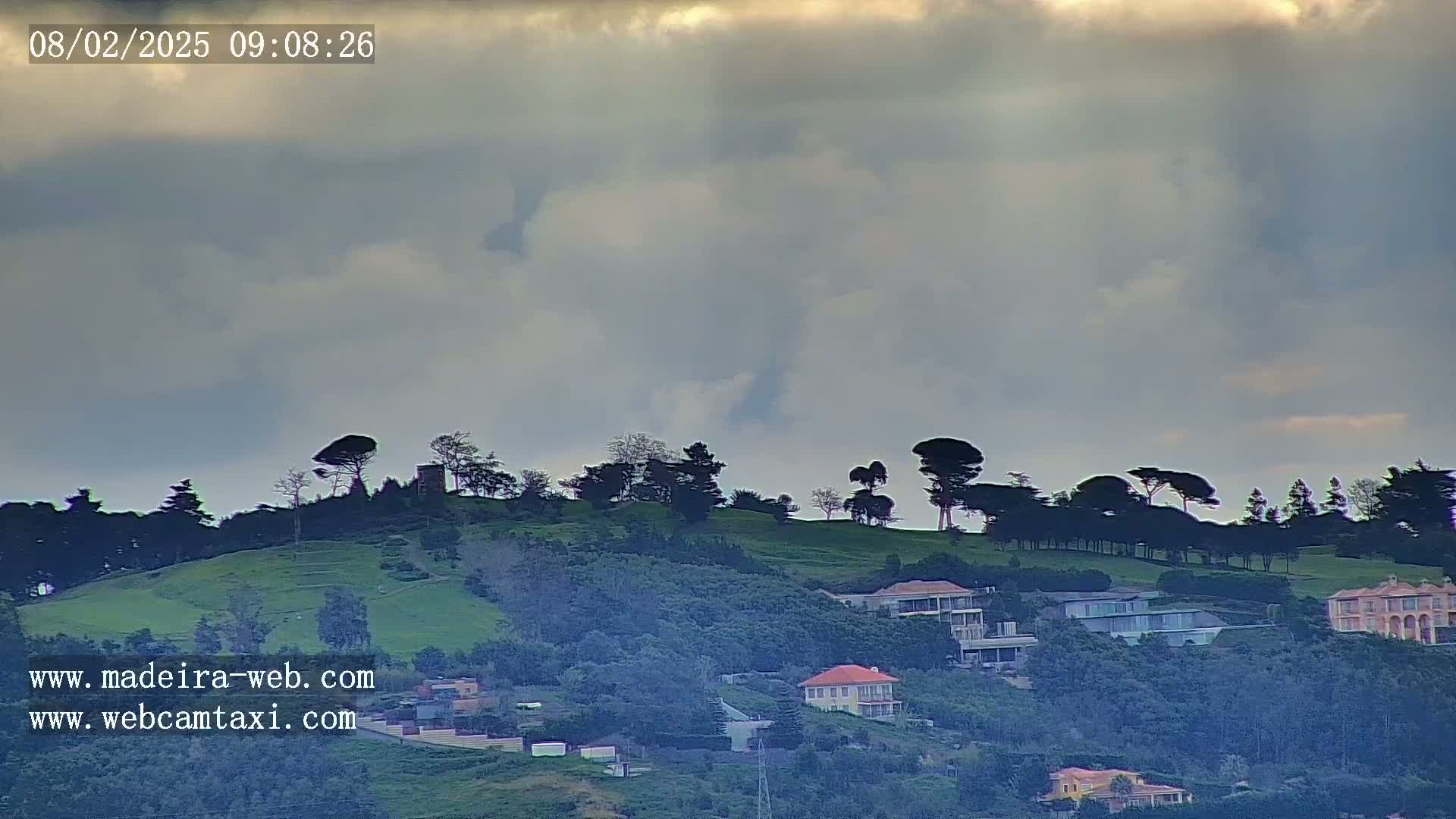 A hill covered in trees and houses is visible under a mostly cloudy sky.