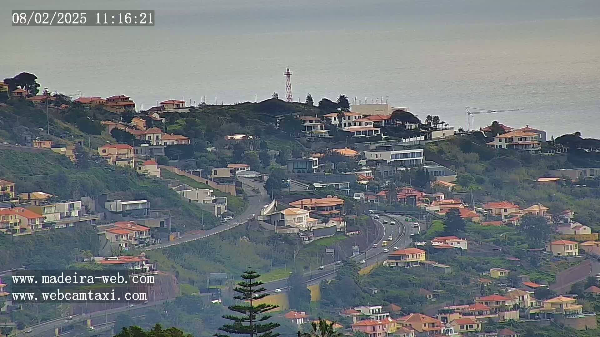 A hillside residential area with numerous houses overlooks a highway and the ocean under an overcast sky.