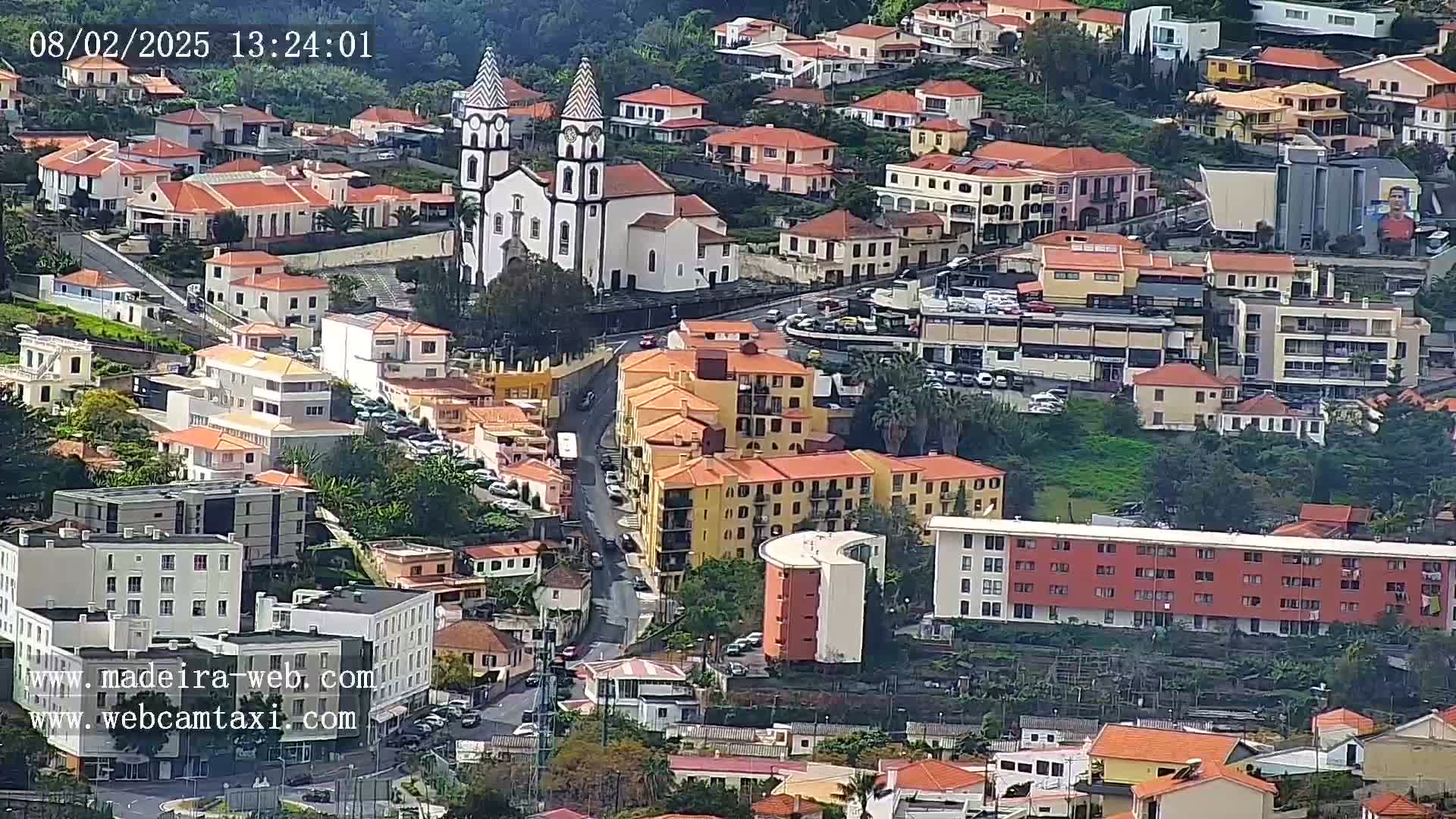 A high-angle view shows a town nestled in a hillside, with numerous buildings of varying sizes and styles, including a prominent white church with twin towers.