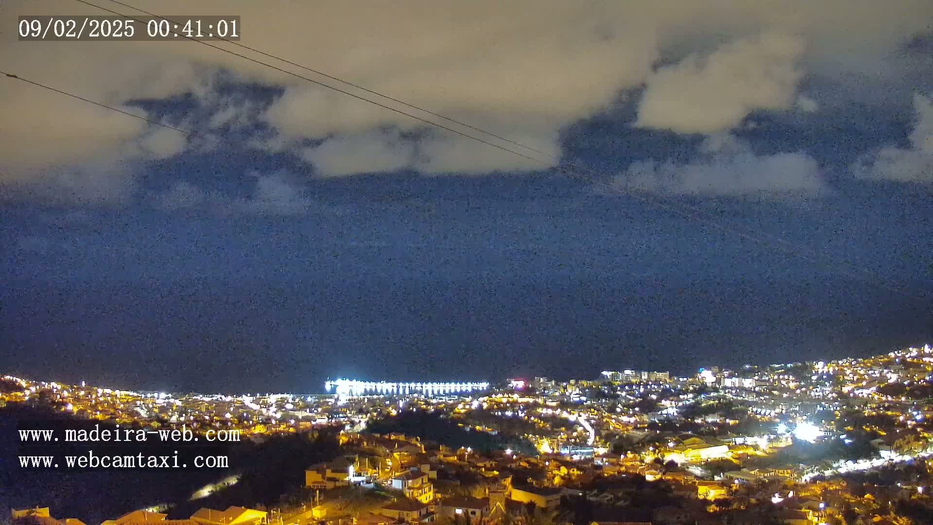 A nighttime view of a city illuminated by lights, with a cloudy sky overhead and a body of water visible in the distance.