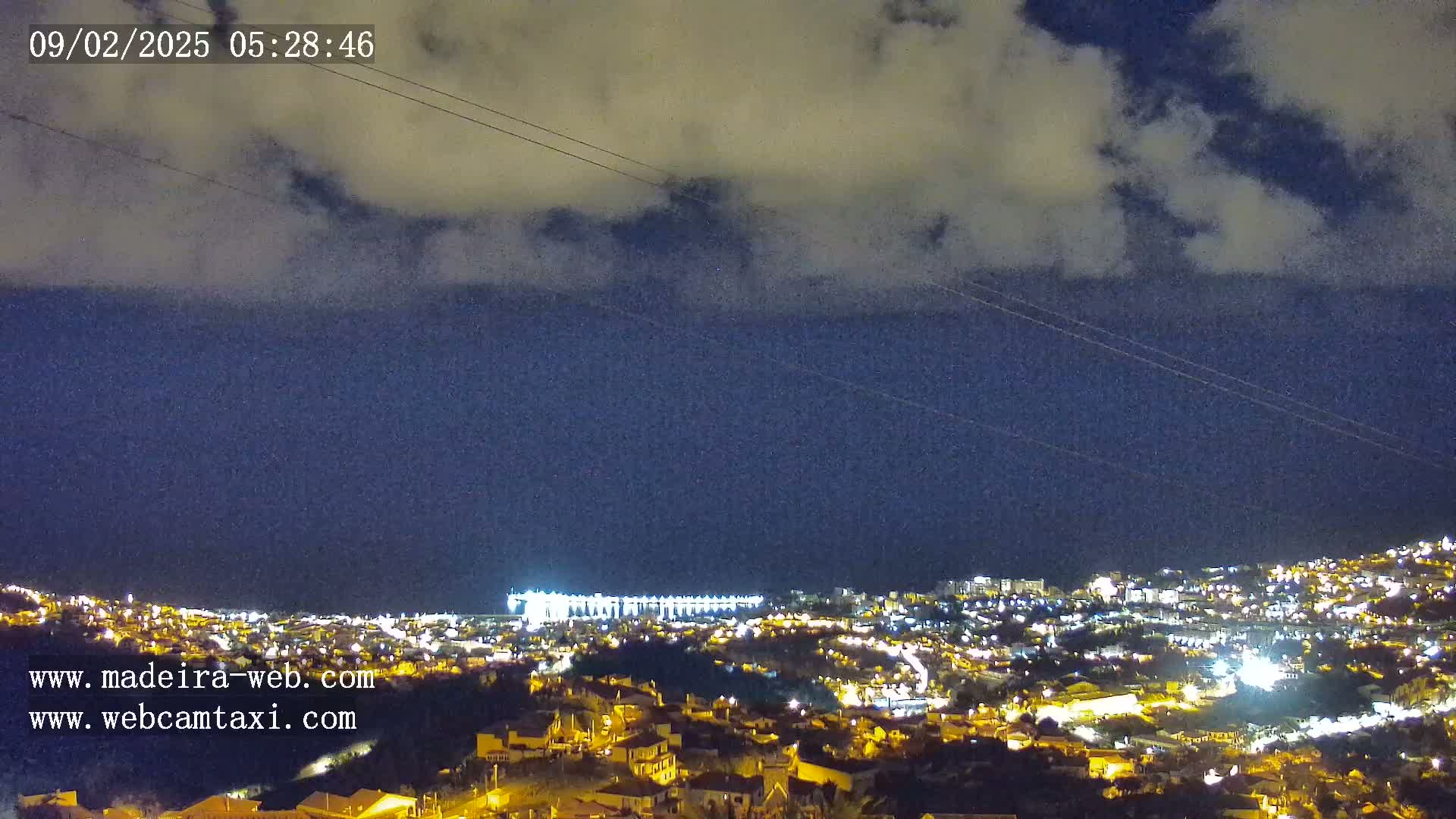 A nighttime view of a coastal town illuminated with many lights under a partly cloudy sky.