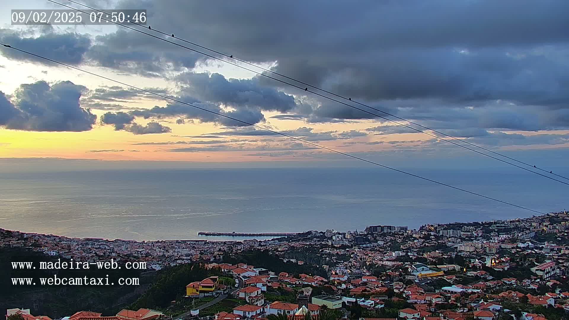 A coastal city at sunset, with a mostly cloudy sky and several birds perched on power lines overhead.