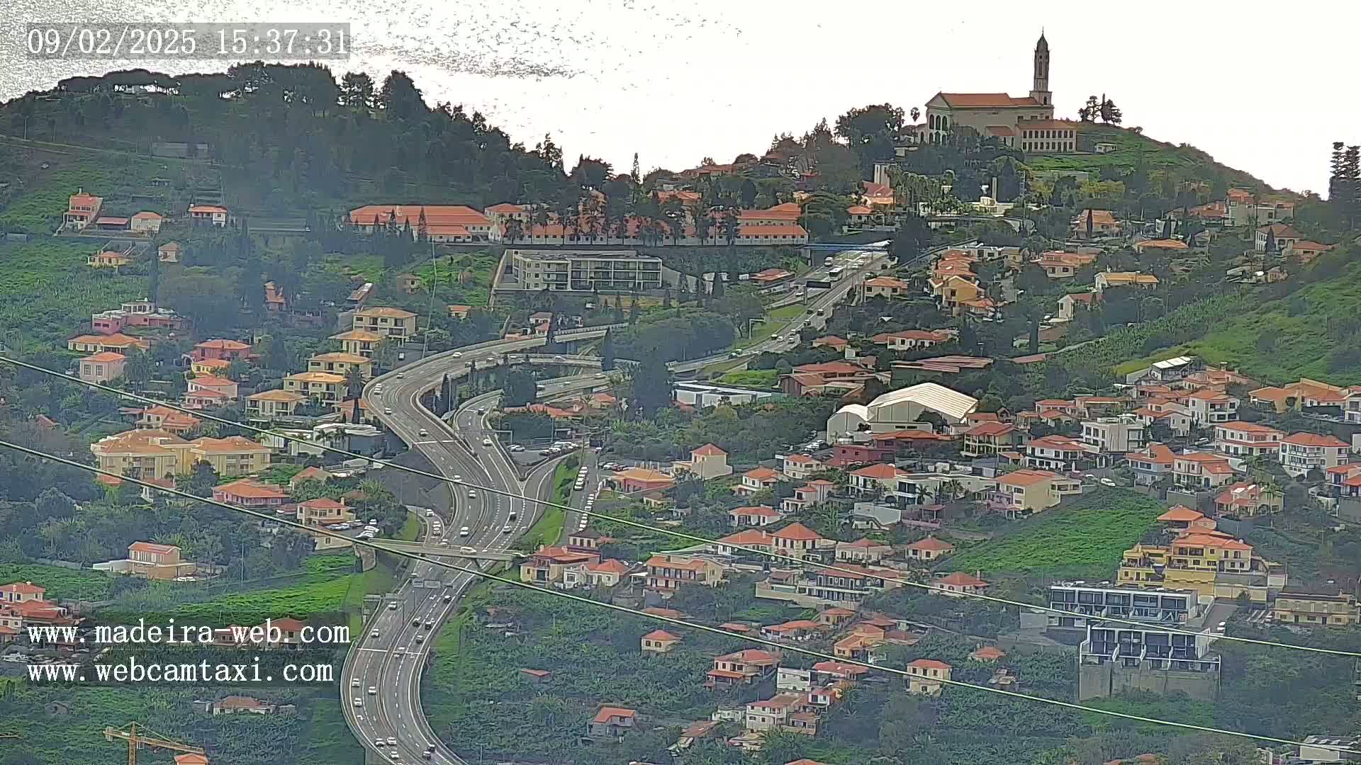 A hillside town with red-roofed houses and a church on a hilltop, overlooked by a multi-lane highway, under an overcast sky.