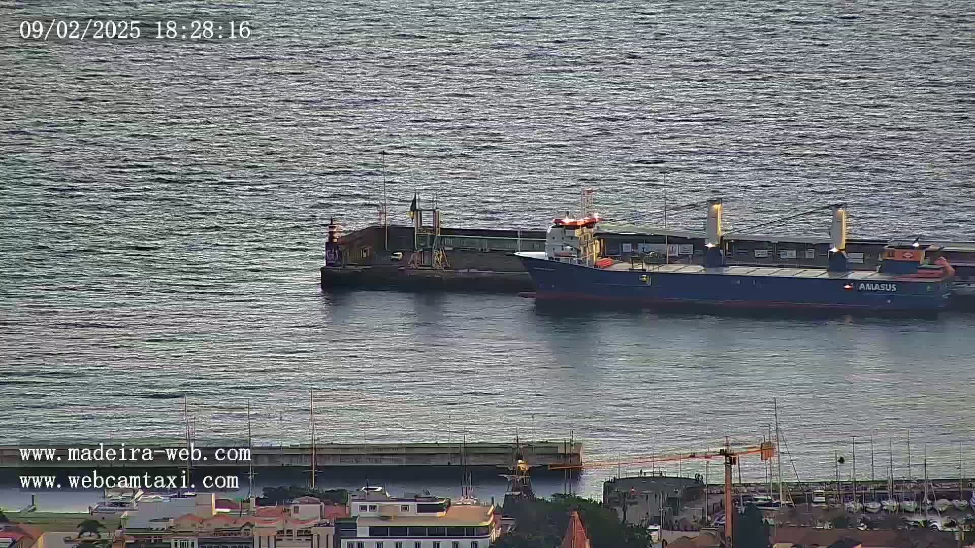 A large cargo ship is docked near a smaller structure in calm water, with a marina and town visible in the foreground.