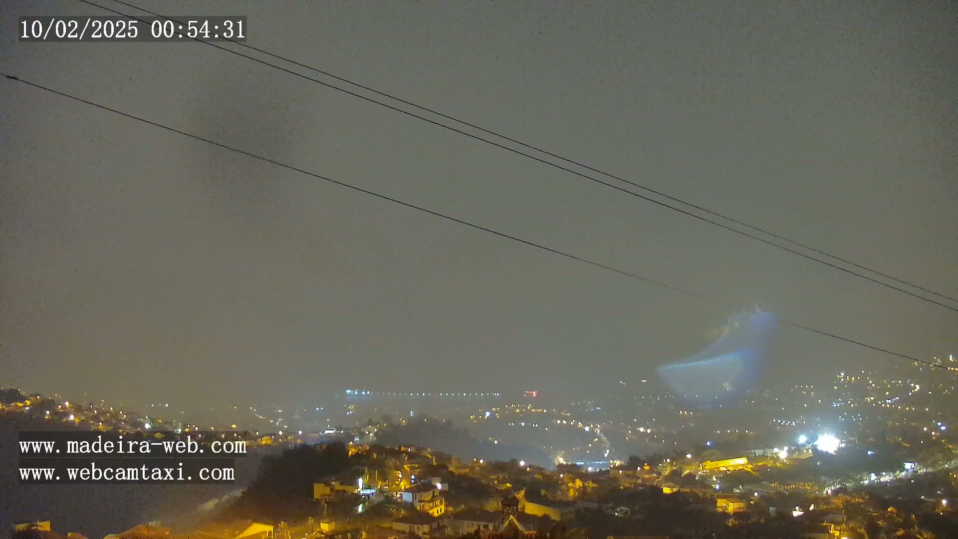A nighttime view of a hazy city illuminated by lights, with power lines visible in the foreground and a faint bluish glow in the distance.