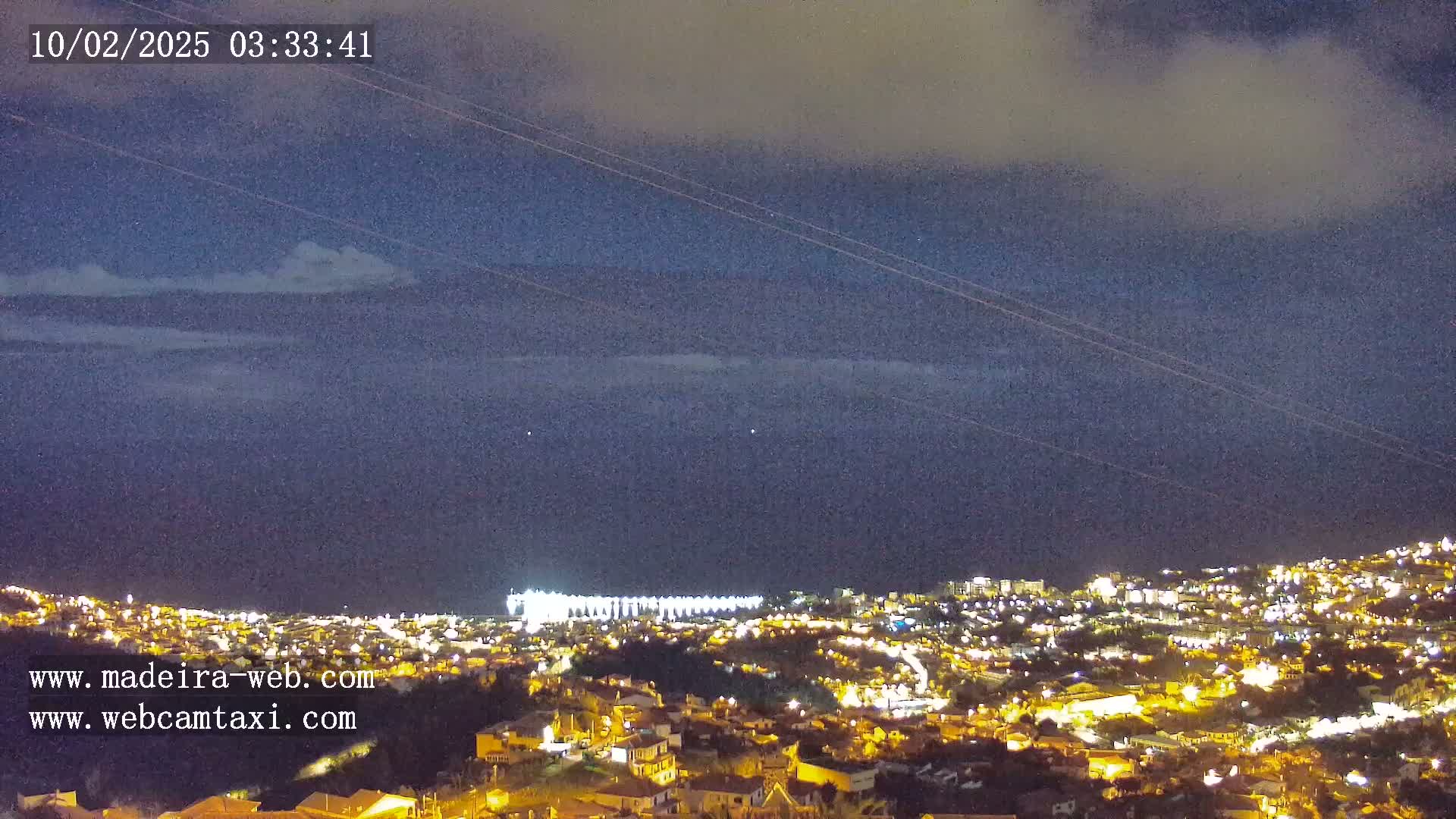 A nighttime view of a coastal city illuminated by lights, under a mostly cloudy sky.