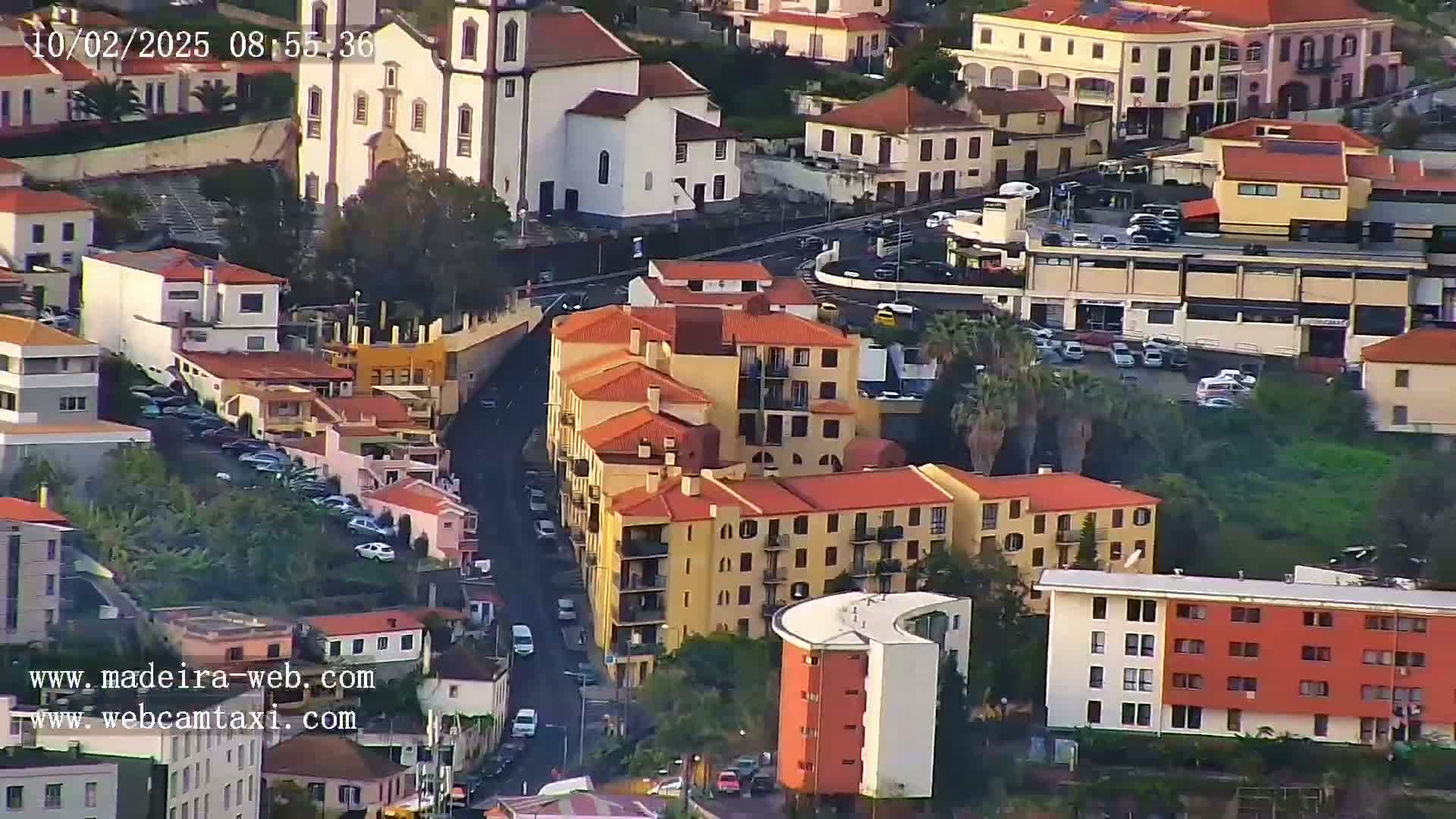 An aerial view shows a city with numerous orange-roofed buildings, a church, and parked cars on a sunny day.