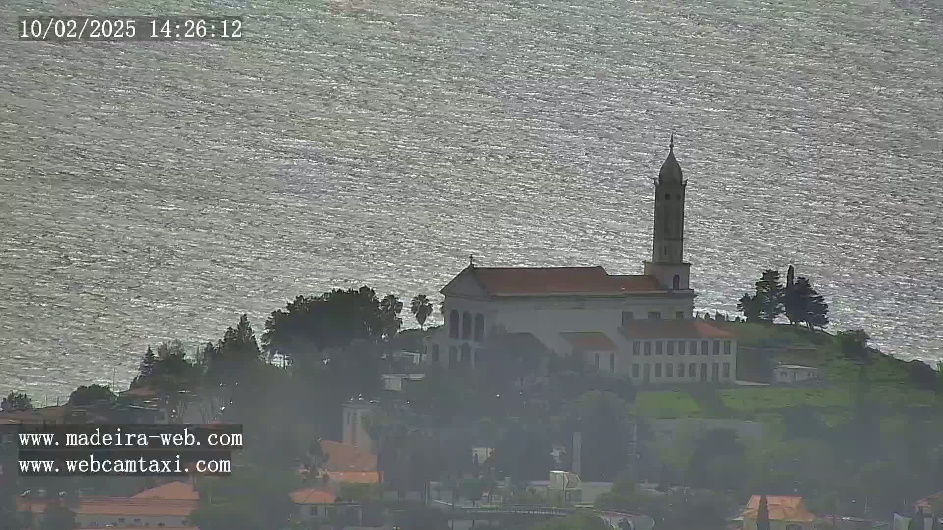 A church with a tall tower sits on a hill overlooking a body of water under somewhat overcast skies.