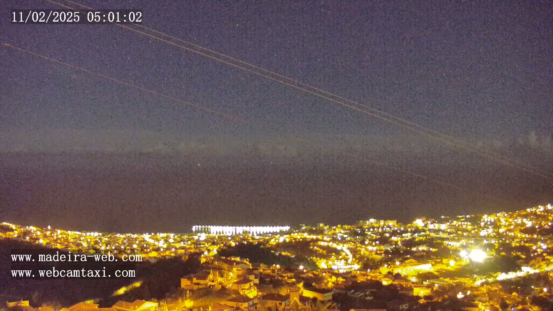 A nighttime, long-range view of a city illuminated by lights against a dark, clear sky with airplane contrails.