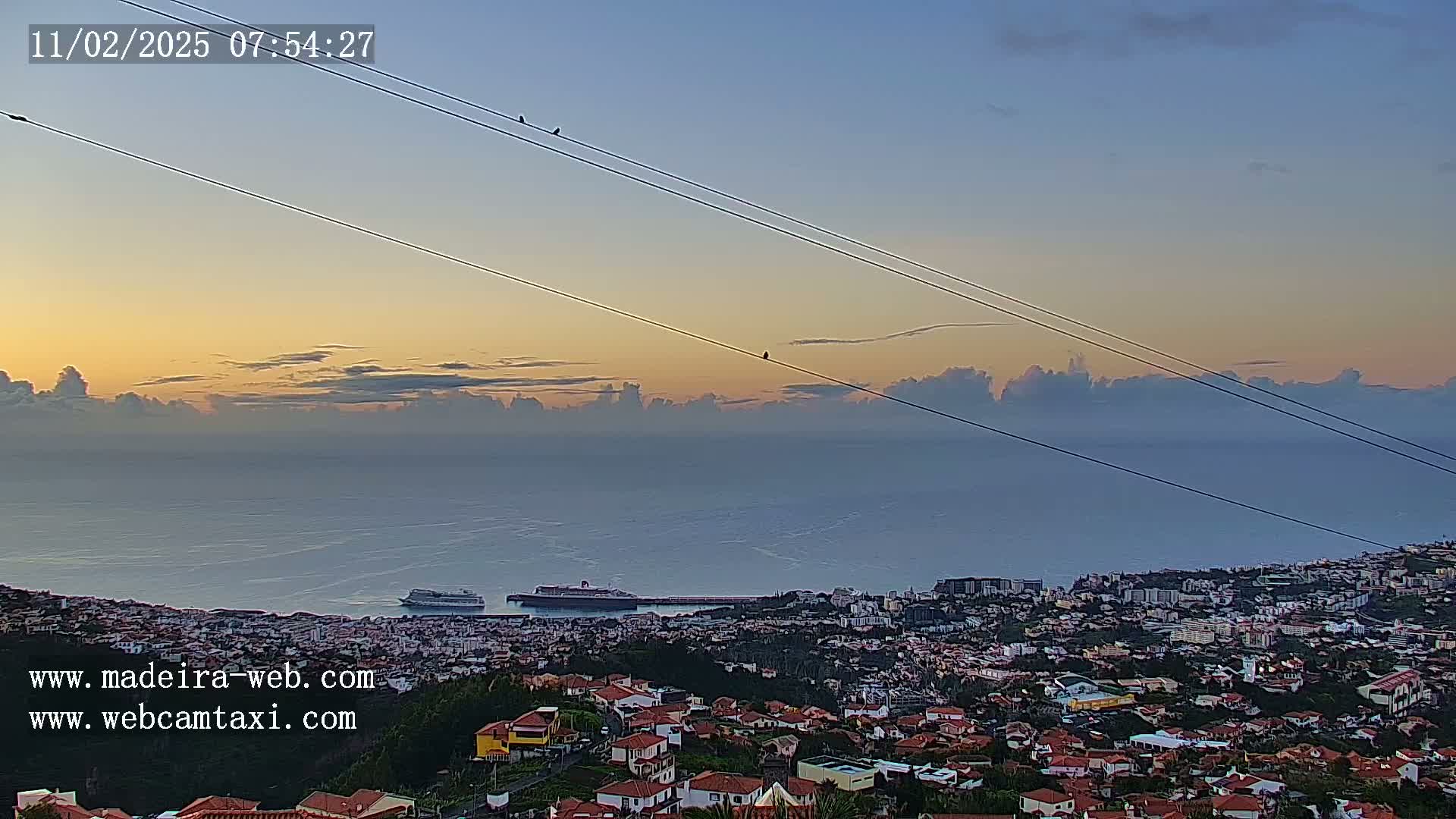 A coastal city at sunrise is viewed from a hillside, with a cruise ship visible in the harbor below and a calm ocean extending to the horizon under a mostly clear sky.