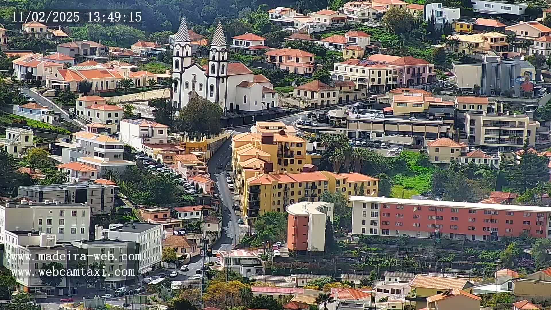 A high-angle view of a town with predominantly red-roofed buildings, a prominent church, and curving streets, on a sunny day.