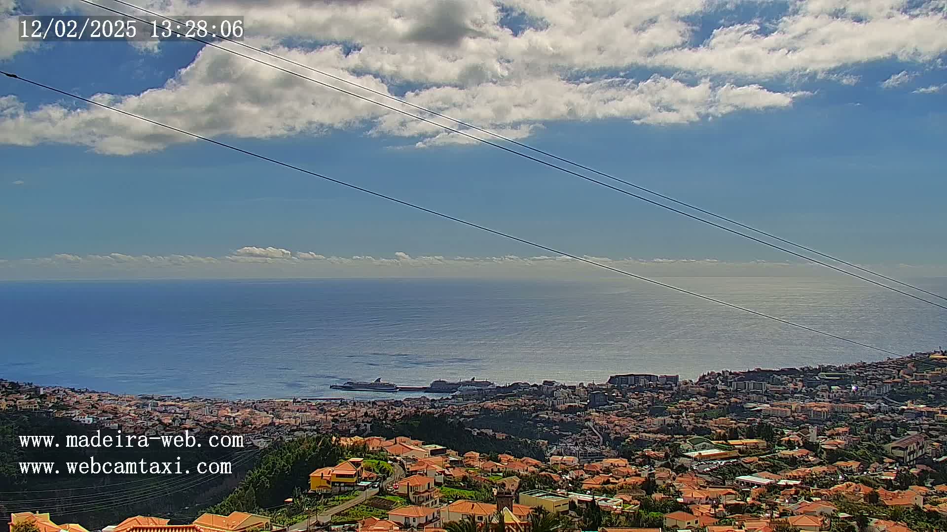 A coastal town with many low-rise buildings is shown under a mostly sunny sky with scattered clouds, overlooking a calm ocean with a few ships visible in the distance.