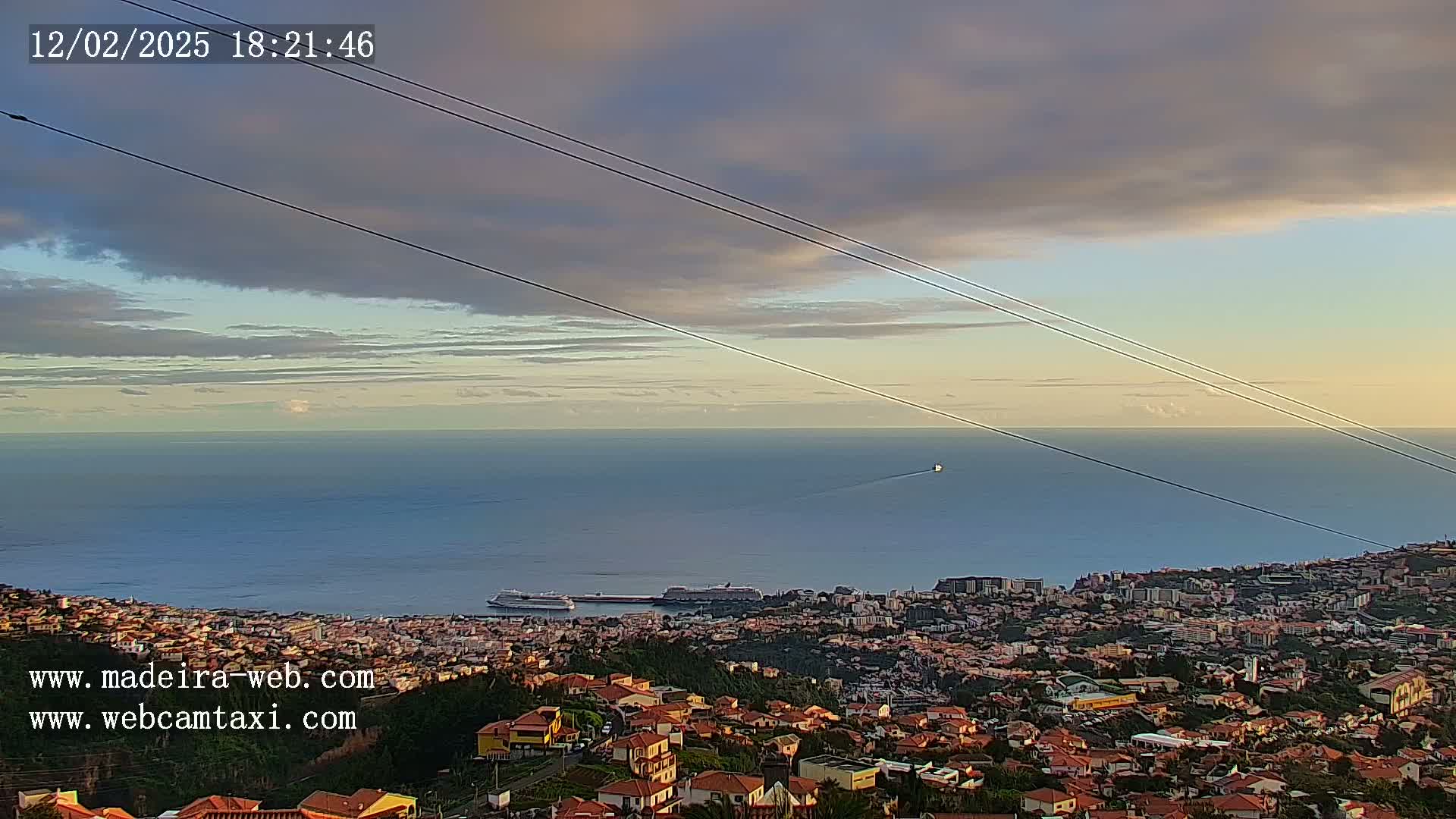 A coastal city with red-roofed houses sits below a calm ocean under a partly cloudy twilight sky.
