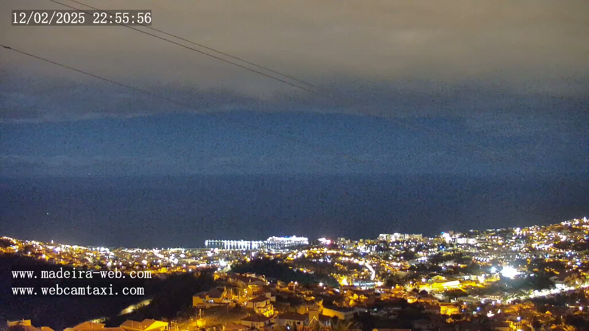 A nighttime view shows a city illuminated against a dark, cloudy sky over a body of water.