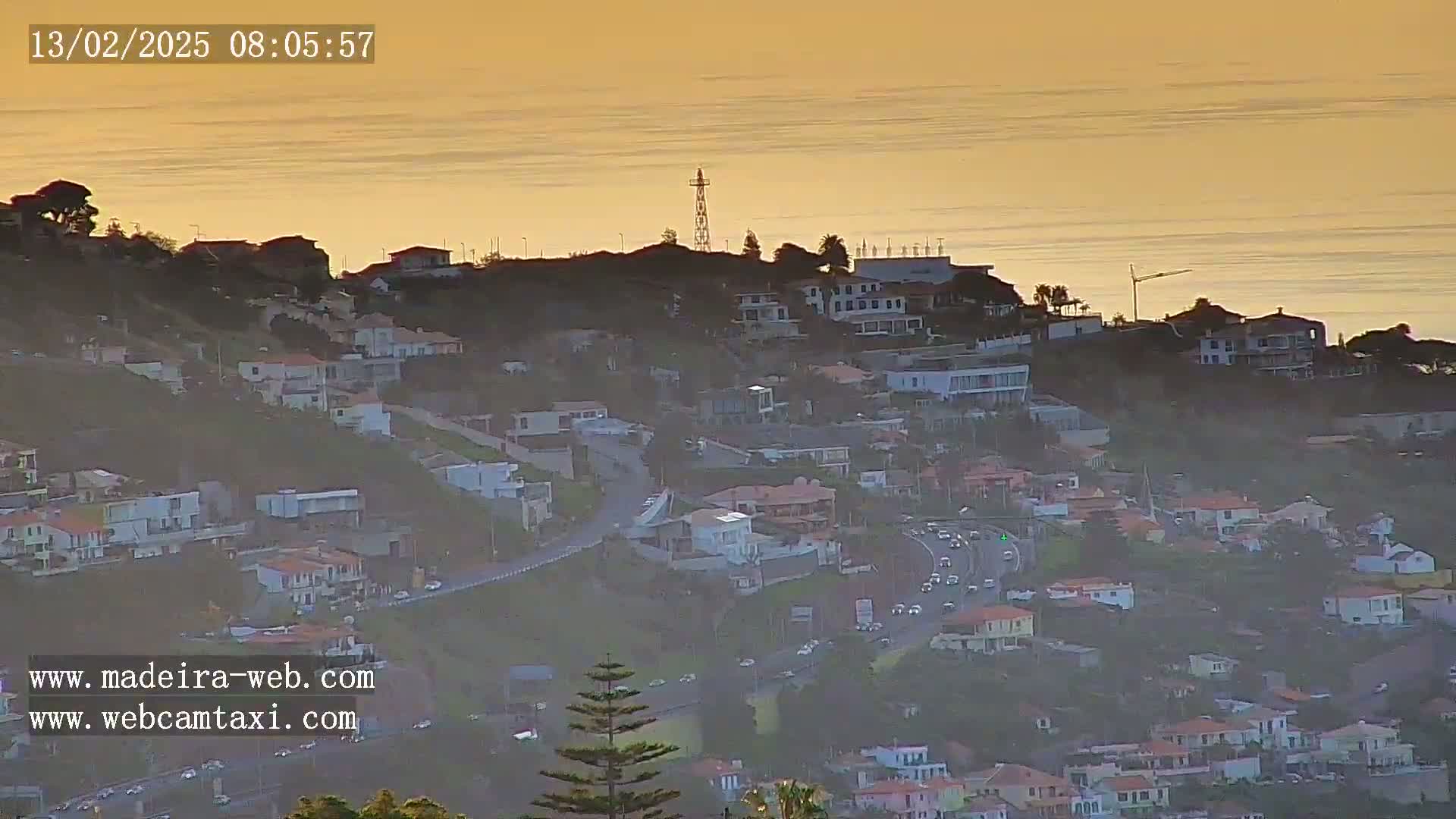 A hillside residential area with a coastal road and ocean view under a hazy, golden-orange sunset.