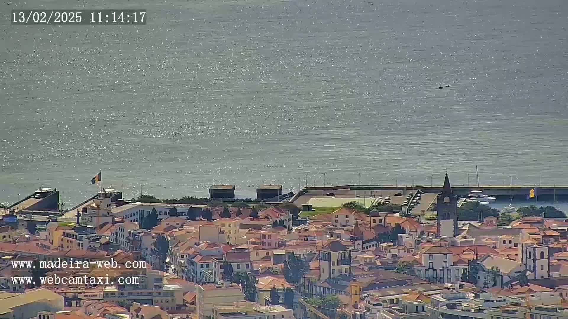 A coastal town with a mix of low-rise buildings and a church steeple is visible, overlooking a calm body of water under a sunny sky.