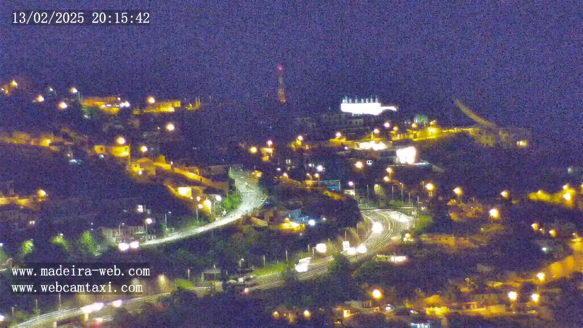 A nighttime, overhead view of a hilly town with roads and illuminated buildings under a dark sky.