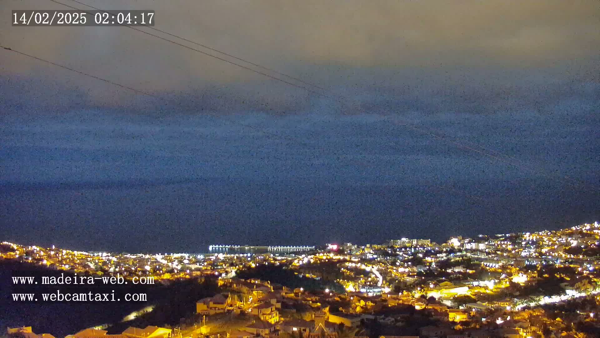 A nighttime view of a coastal town illuminated by lights, with a dark, cloudy sky and calm ocean extending to the horizon.