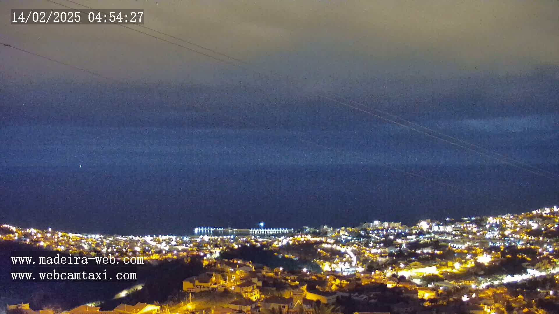 A nighttime view of a coastal town illuminated by lights, with a dark blue sea under a mostly clear, dusky sky.