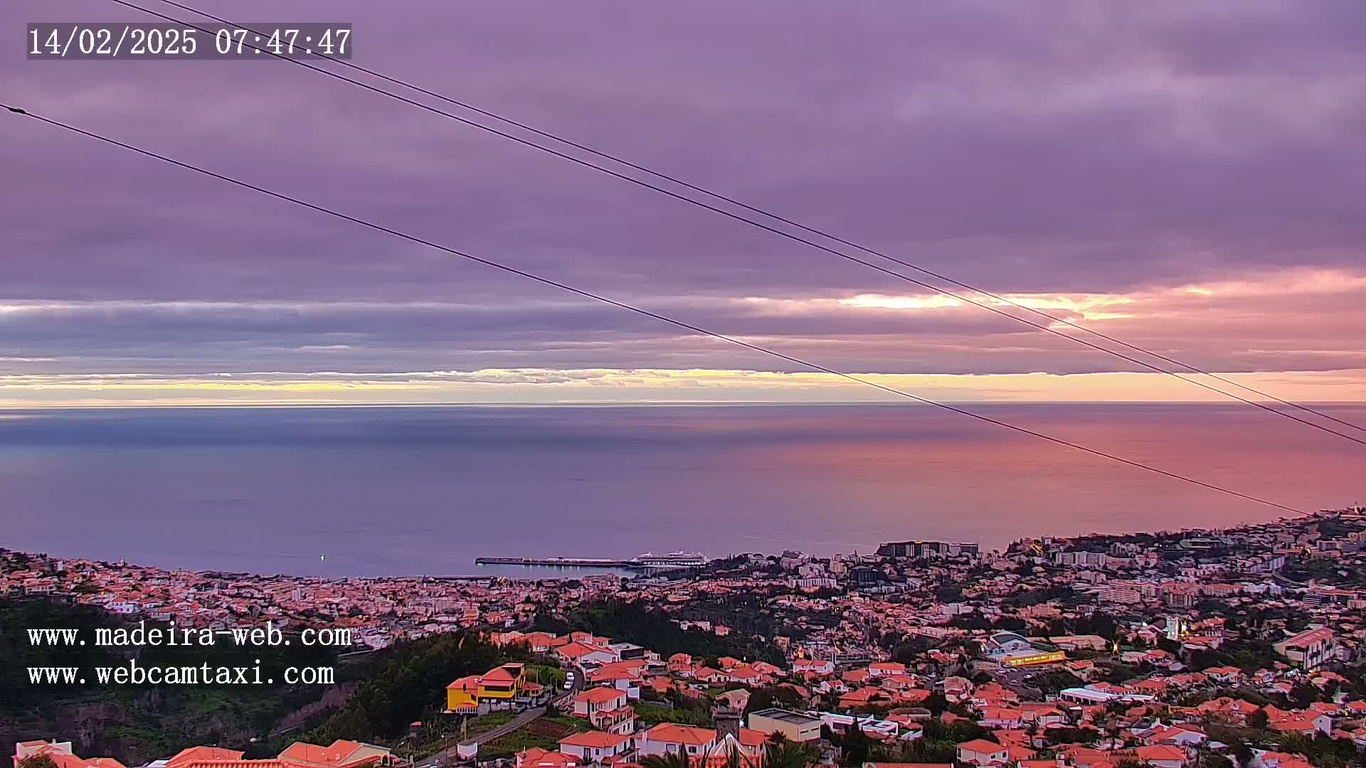 A coastal city with red-tiled roofs stretches below a serene sunset over a calm ocean under a purple and pink sky.
