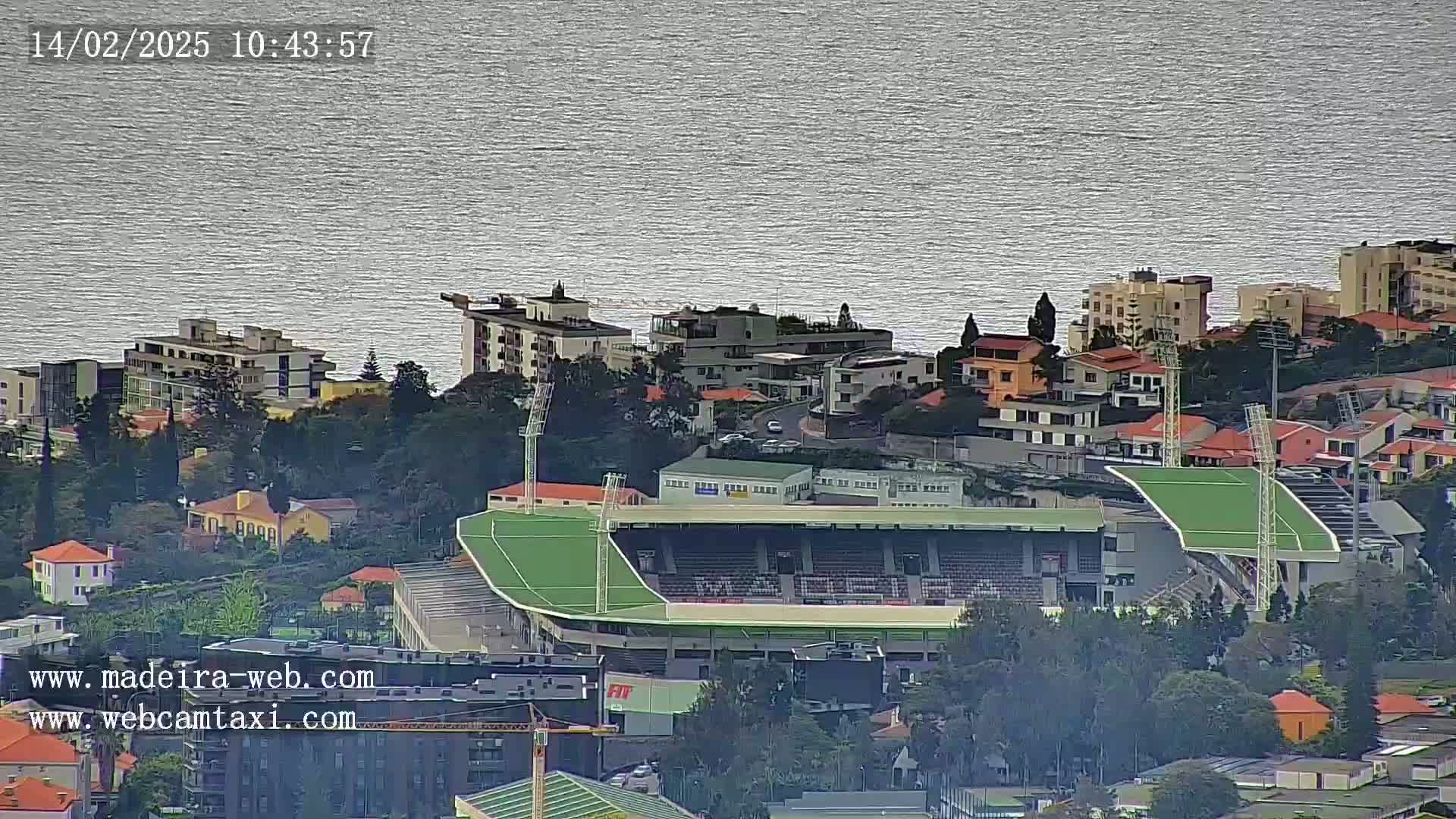 An elevated view shows a soccer stadium with a green field nestled among residential buildings near a calm body of water under a cloudy sky.