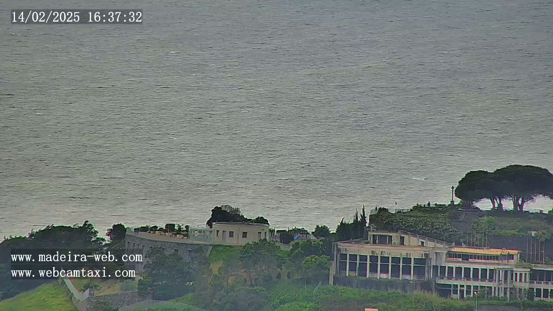 A calm, gray ocean stretches to the horizon, overlooking a coastal area with buildings and trees on a slightly overcast day.