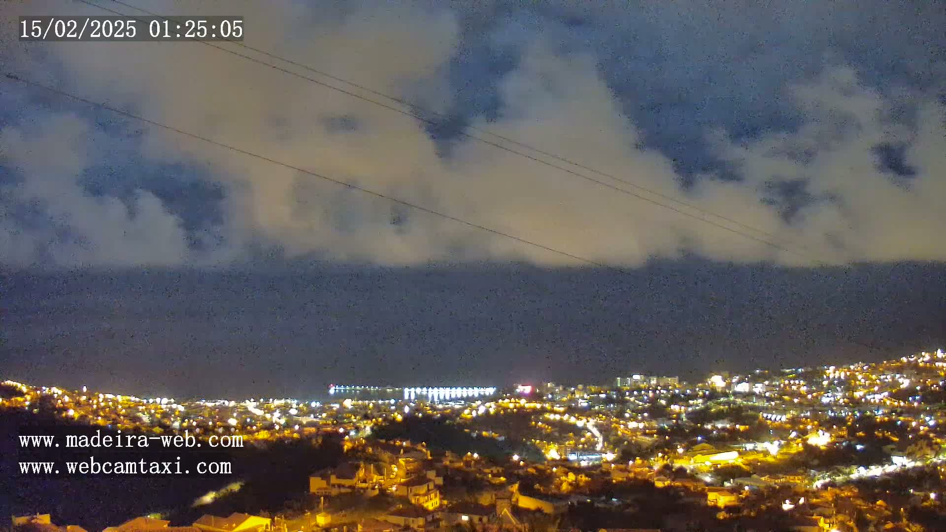 A nighttime view of a coastal city illuminated with lights under a partly cloudy sky.