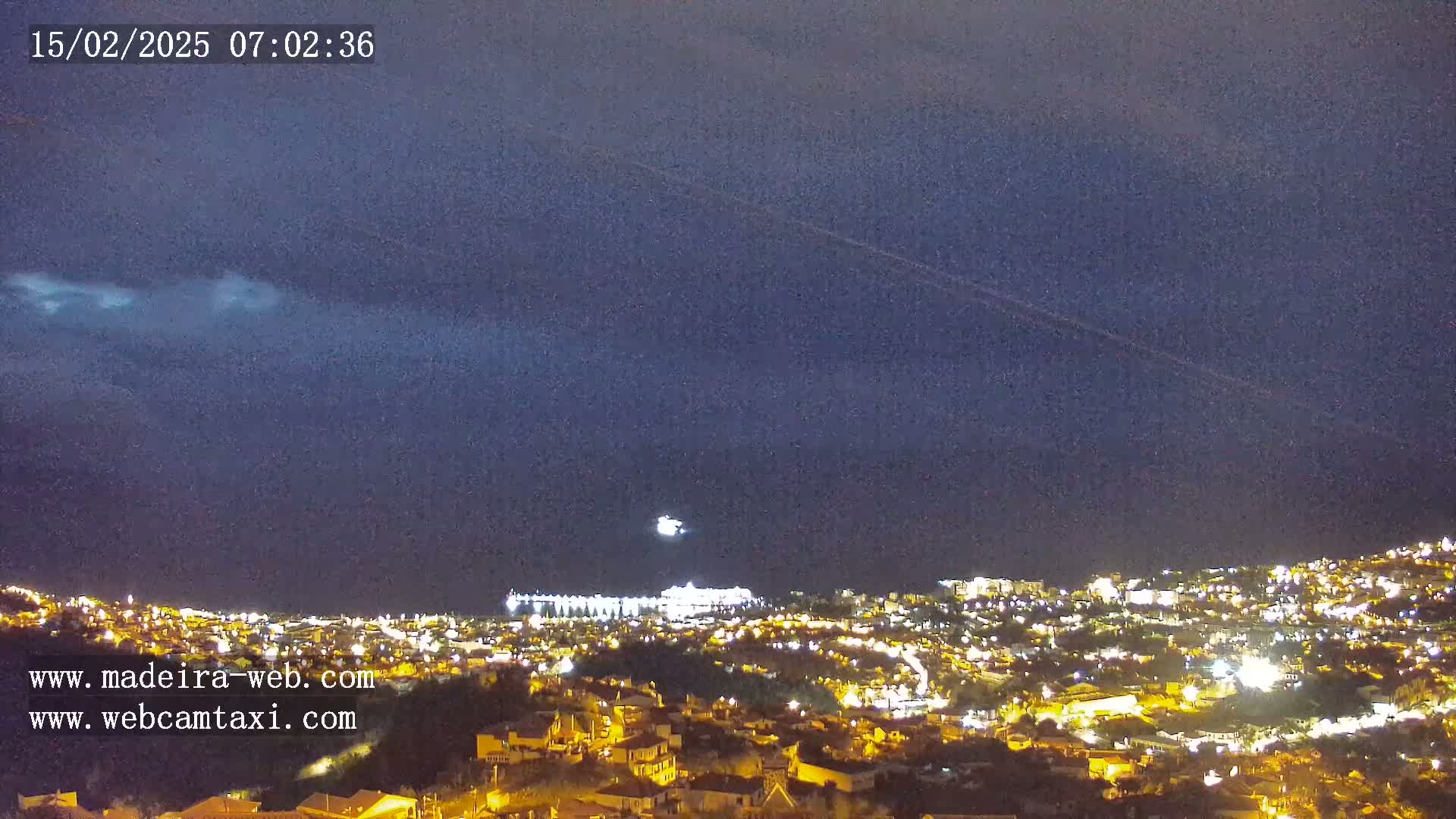 A nighttime view of a city illuminated by lights under a mostly clear, dark blue sky with a few clouds and a bright moon.