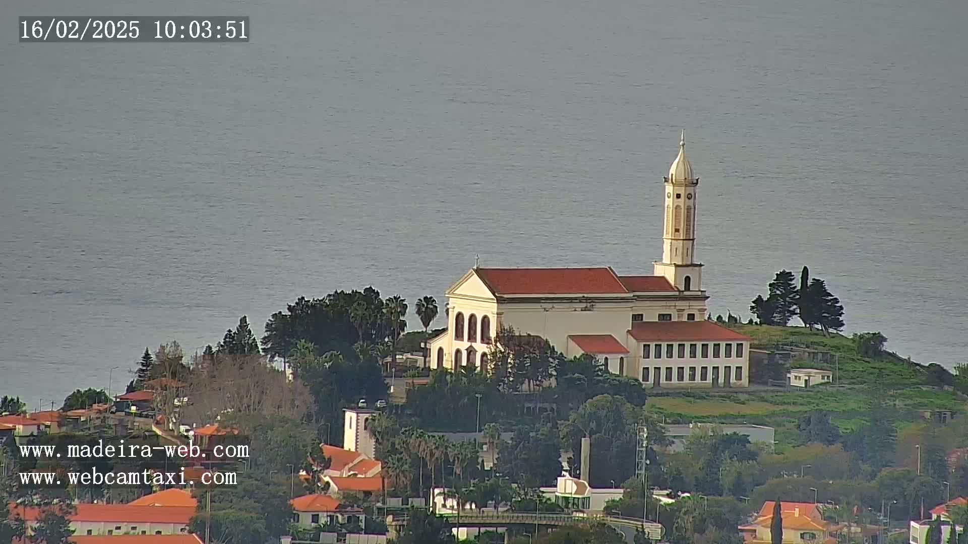 A church with a tall steeple sits on a small hill overlooking a calm body of water on a somewhat overcast day.