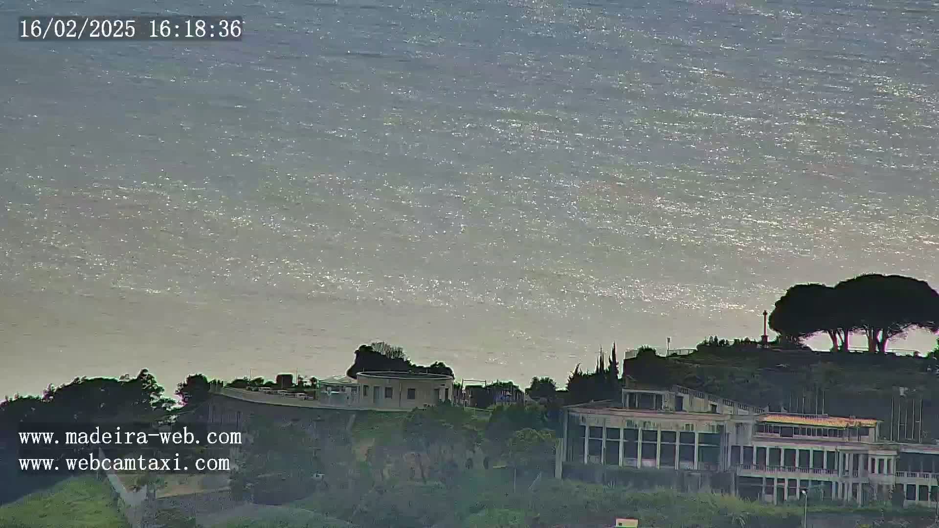 A calm, glittering ocean stretches to the horizon behind a hilltop featuring buildings and a few dark green trees.