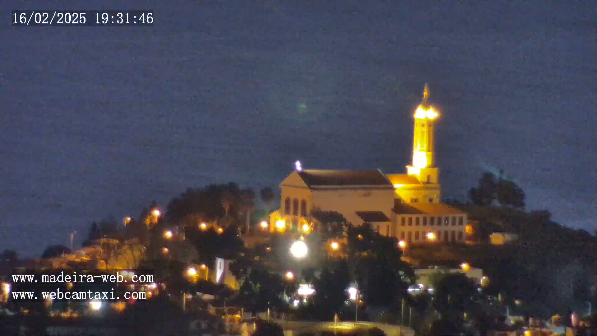 A brightly lit church sits atop a hill at night, overlooking a dark body of water under a clear, dark sky.