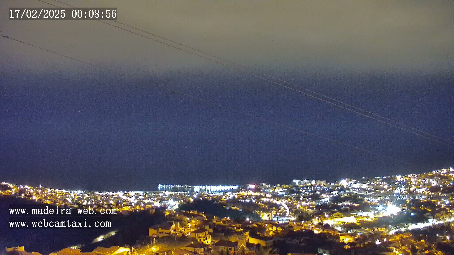 A nighttime view of a city illuminated by lights stretches to a dark ocean under a mostly clear, dark blue sky.