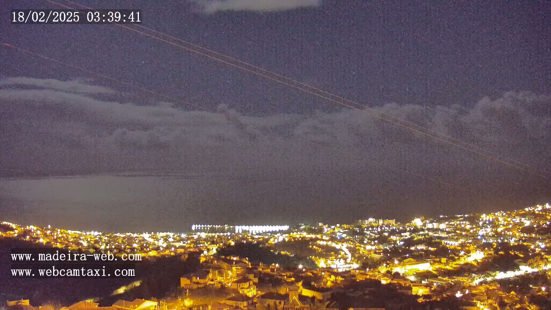 A nighttime view of a coastal town illuminated by lights, under a dark sky with thin clouds.