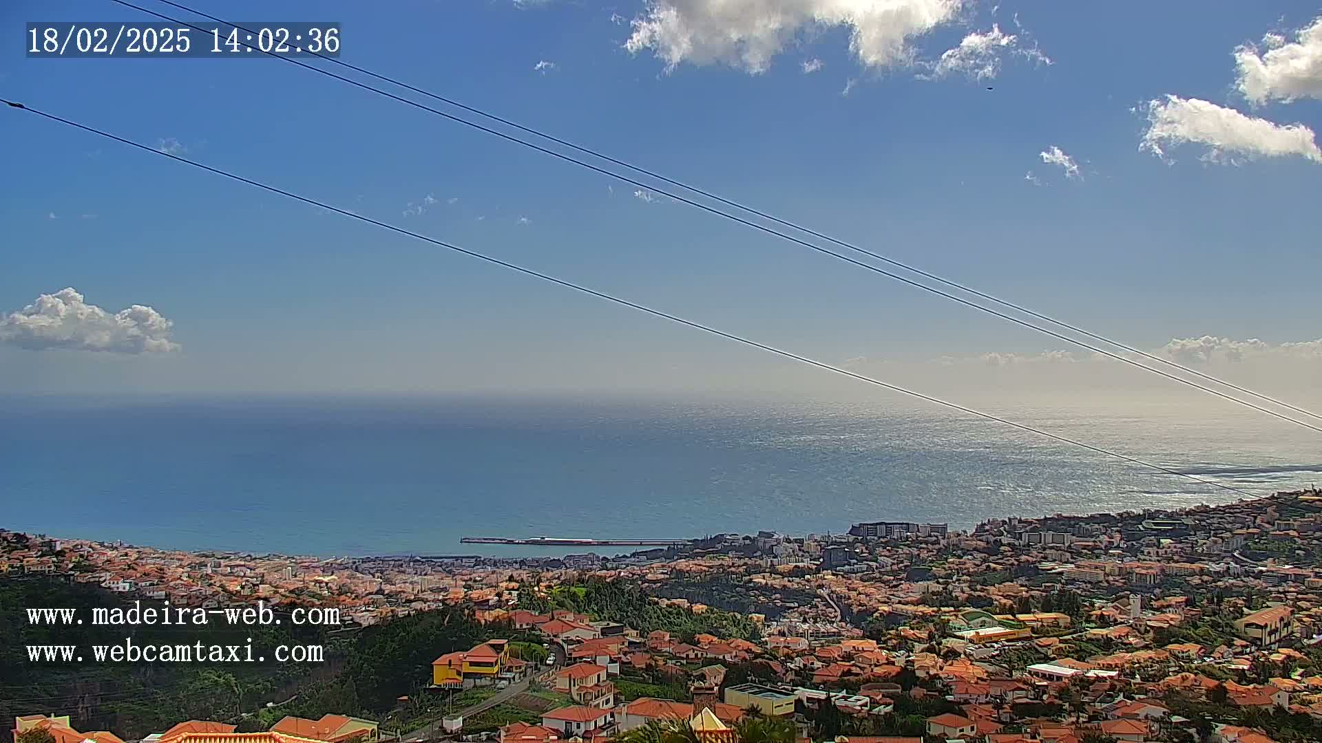 A coastal town with red-roofed buildings sprawls below a clear blue sky with a few scattered clouds, overlooking a calm ocean.