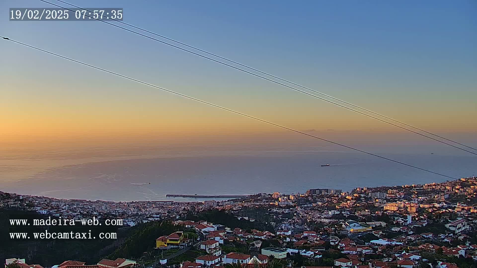 A coastal city at sunset is visible from a hilltop, with calm ocean waters extending to the horizon under a clear sky.